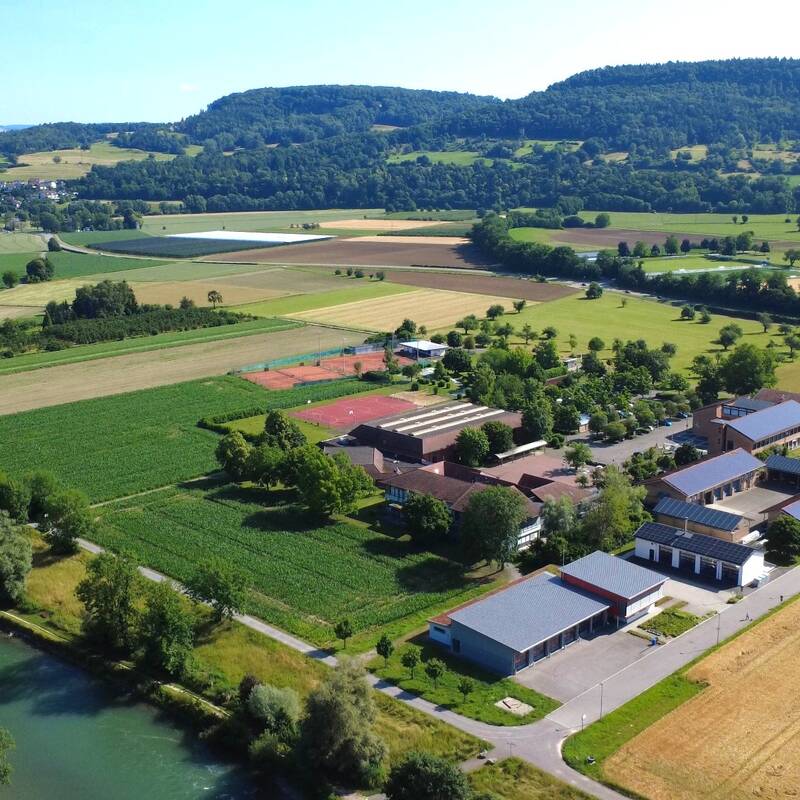 Eine Luftaufnahme zeigt eine grüne, ländliche Landschaft mit Feldern, einem Fluss und Gebäuden, umgeben von Hügeln und vereinzelten Bäumen.