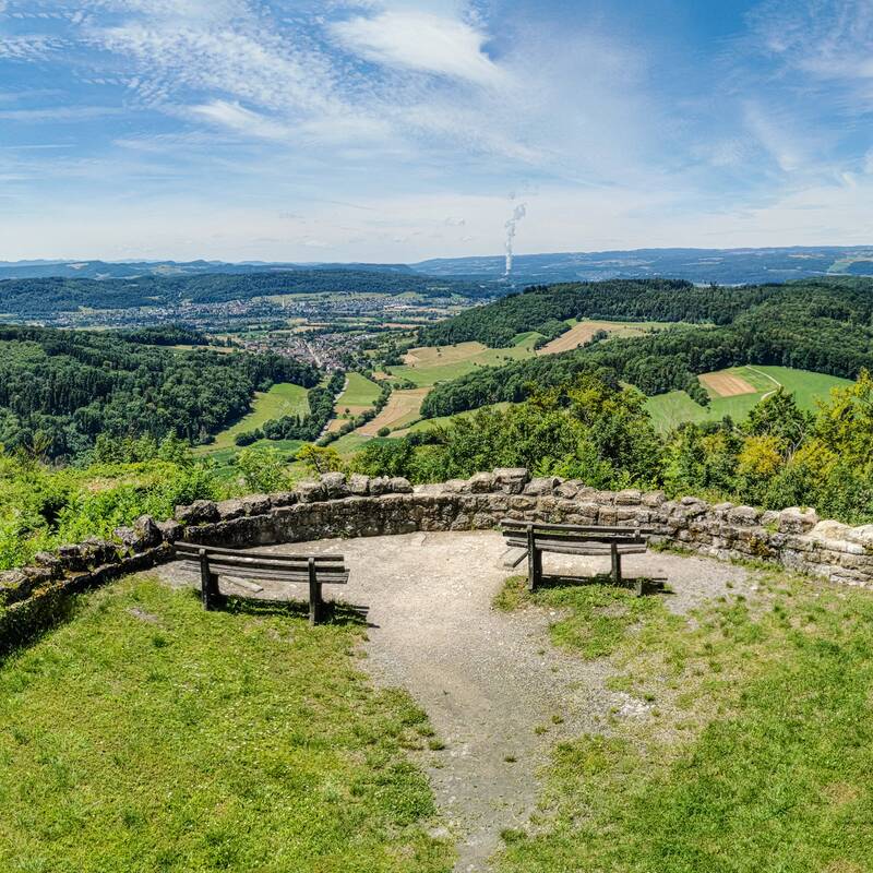 Panoramablick auf eine hügelige Landschaft mit Wäldern und Feldern, betrachtet von einem Aussichtspunkt mit einer hölzernen Bank und Steinmauer.