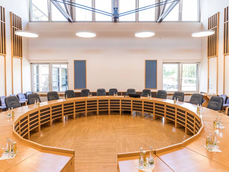 Ein heller, moderner Konferenzsaal mit einer ovalen Sitzordnung aus Holz und blauen Stühlen, umgeben von großen Fenstern und hohen Decken.