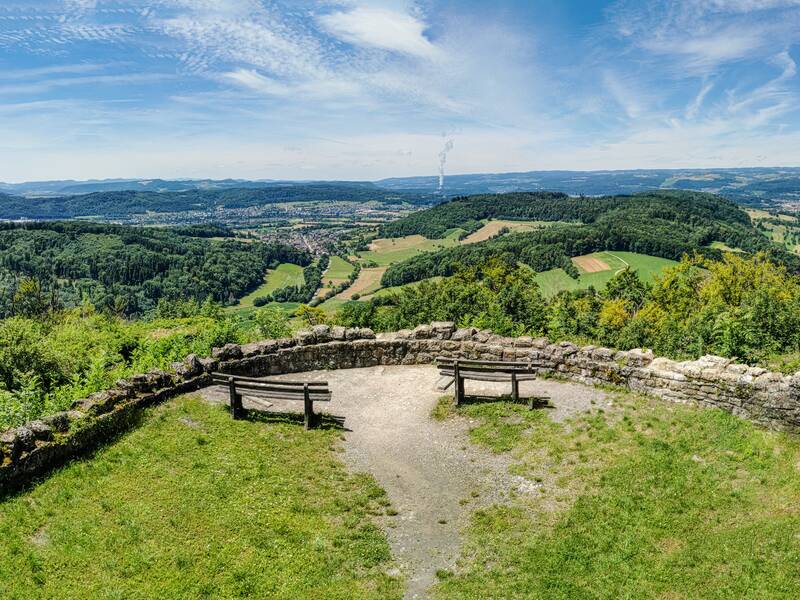 Eine steinerne Aussichtsplattform mit einer Bank bietet einen weiten Blick über eine grüne, hügelige Landschaft unter einem blauen Himmel mit vereinzelten Wolken.