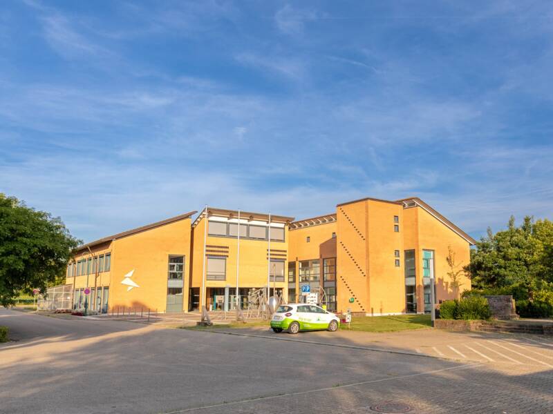 Eine breite Straße führt zu einem modernen Gebäudekomplex unter einem blauen Himmel, begleitet von einem grün-weißen Auto und umgeben von kleinen Bäumen.