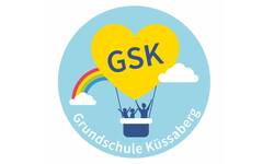 Ein gelbes Herz mit der Aufschrift "GSK" schwebt als Heißluftballon über Wolken, getragen von einem Regenbogen, während zwei kindliche Silhouetten in der Gondel jubeln, unterhalb steht "Grundschule Küssaberg".