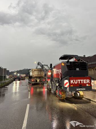 Eine Baustelle bei schlechtem Wetter, auf der Fahrzeuge auf nasser Straße arbeiten, darunter eine große, rot-graue Baumaschine mit dem Schriftzug "KUTTER".