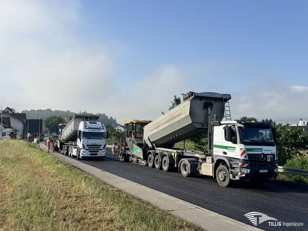 Auf einer Landstraße wird Straßenbauarbeit durchgeführt, bei der mehrere Lastwagen und Bauarbeiter beteiligt sind.