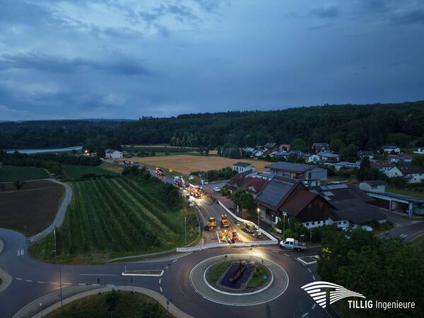 Eine nächtliche Luftaufnahme eines Kreisverkehrs zeigt eine Straßenbaustelle mit beleuchteten Fahrzeugen, umgeben von Feldern und Häusern.