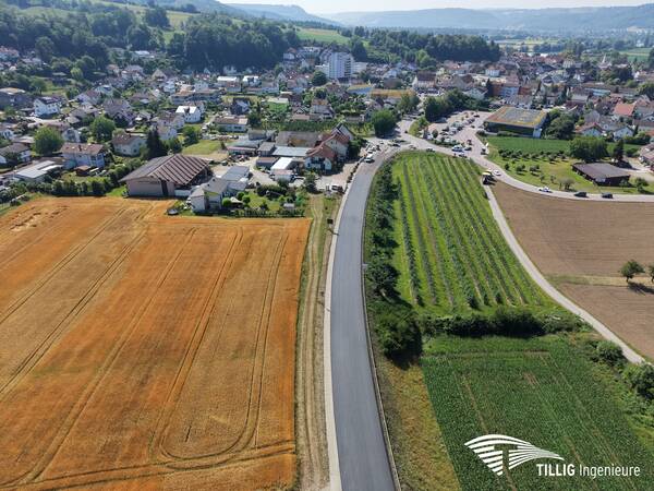 Die Aufnahme zeigt eine ländliche Landschaft mit einem neu befestigten Weg, der zwischen landwirtschaftlichen Feldern und einem Dorf mit zahlreichen Gebäuden verläuft.