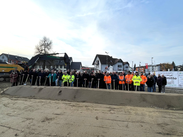 Eine Gruppe von Menschen steht mit Schaufeln vor einem Erdwall auf einer Baustelle, im Hintergrund sind Gebäude und ein Bagger zu sehen.