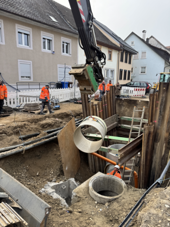 Eine Baustelle mit mehreren Bauarbeitern in orangefarbener Schutzkleidung, die an einem Rohrgraben in einer Wohngegend arbeiten, während ein Kran ein großes Rohrteil bewegt.