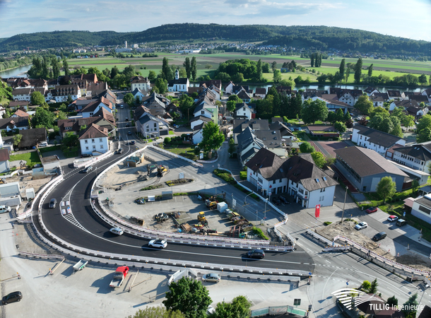 Eine Luftaufnahme zeigt eine Stadtlandschaft mit einem im Bau befindlichen Straßenkreisel, umgeben von Wohnhäusern und grünen Feldern im Hintergrund.