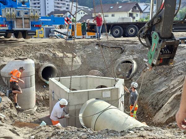 Auf einer Baustelle wird ein großes Betonbauteil mit einem Kran in einen ausgehobenen Graben gehoben, während Arbeiter bei der Platzierung helfen.