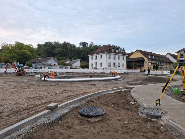 Eine Baustelle mit Arbeitern, die an einem Kreisverkehr in einer kleinen Stadt arbeiten, umgeben von Bäumen und traditionellen Gebäuden.