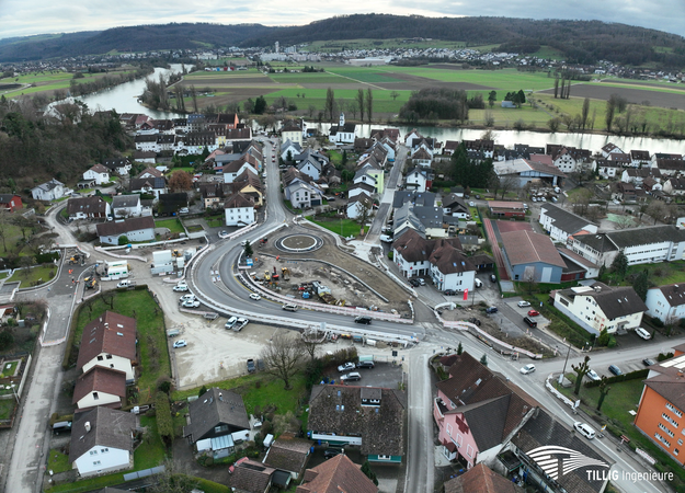 Luftaufnahme einer kleinen Stadt mit einem neu errichteten Kreisverkehr in der Mitte und einer angrenzenden Flusslandschaft im Hintergrund.