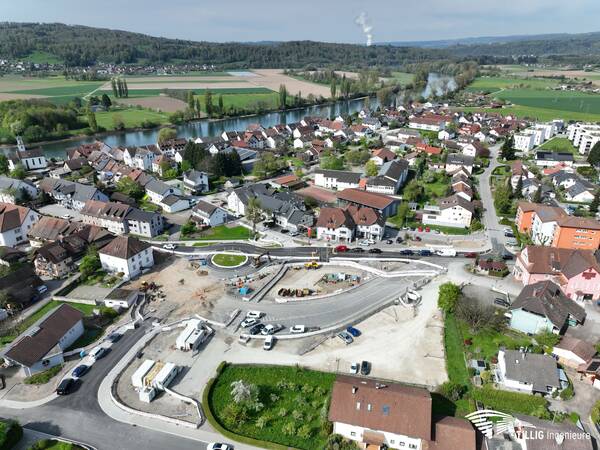 Das Bild zeigt eine Luftaufnahme einer Kleinstadt mit einem Fluss im Hintergrund, wobei im Vordergrund eine Baustelle zu sehen ist, die von zahlreichen Gebäuden umgeben ist.
