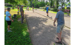 Mehrere Personen, meistens scheinbar im Seniorenalter, spielen Boule in einem Park mit Bäumen und Bänken im Hintergrund.