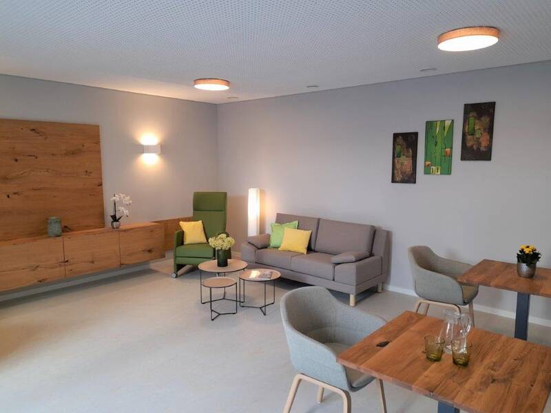 Ein gemütlicher, heller mit Licht durchfluteter, modern eingerichteter Raum mit grauem Sofa, grüner Polsterung, Holzelementen, Bildern an der Wand und einem kleinen Tischbereich.