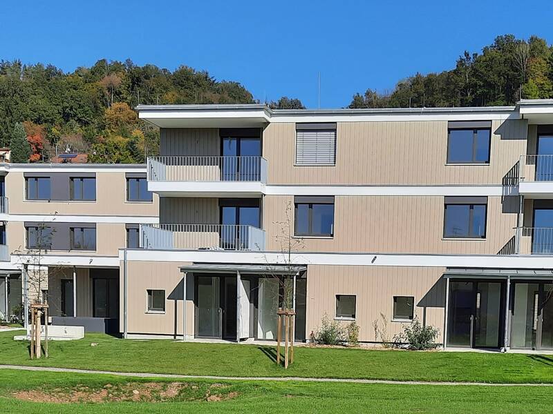 Das Bild zeigt ein modernes dreigeschossiges Wohngebäude mit Balkonen und großen Glasfenstern, umgeben von grünen Rasenflächen und Bäumen im Hintergrund.