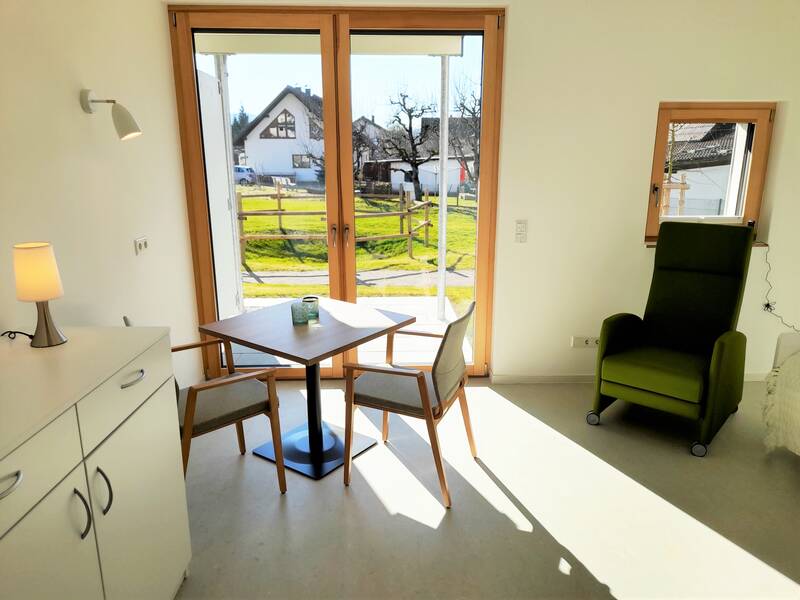 Ein schlicht eingerichtetes, helles Zimmer mit einem kleinen Tisch und zwei Stühlen vor einer großen Fensterfront, durch die ein sonniger Garten zu sehen ist. Rechts im Raum ein grüner Sessel.