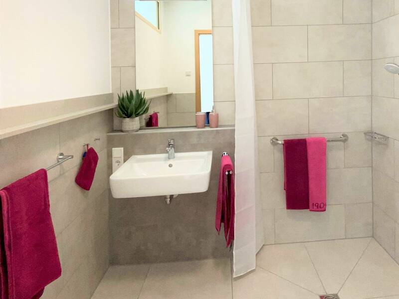 Ein helles, barrierefreies Badezimmer mit weißer Keramik, beigefarbenen Fliesen, pinken Handtüchern und einer Pflanze auf dem Waschbecken.