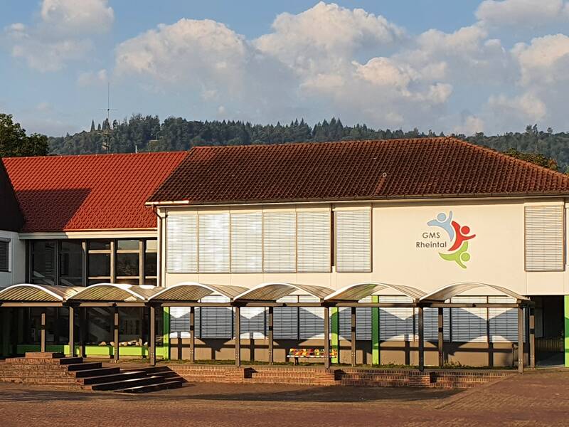 Das Foto zeigt ein modernes Schulgebäude mit rotem Ziegeldach und einem Logo, das auf der Fassade angebracht ist, umgeben von Bäumen und einem blauen Himmel im Hintergrund. Im Vordergrund der Schulhofplatz mit einem überdachten Weg zum Eingang.  In der Mitte des Weges steht eine Sitzbank. 