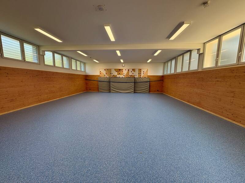 Ein leerer Raum mit blauen Bodenbelag, Holzvertäfelung an den Wänden, großen Fenstern und einer Kletterwand mit Matten auf der Rückseite.