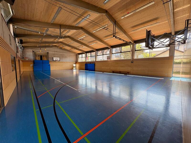 Eine leere Sporthalle mit blauen Wänden und einem Spielfeld auf dem Boden, beleuchtet durch große Fenster an der rechten Seite.