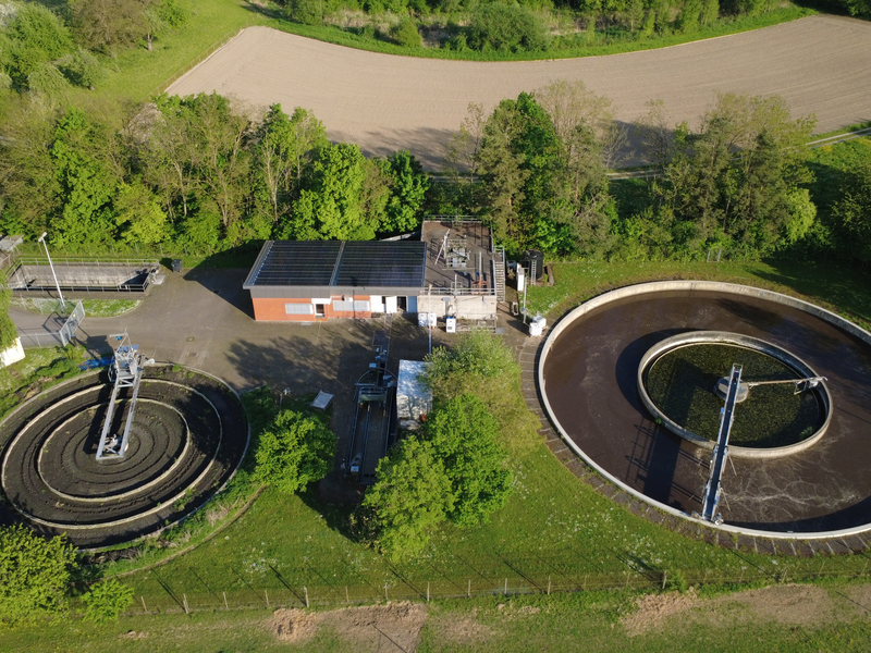 Luftaufnahme einer Kläranlage mit zwei runden Becken, umgeben von grünen Bäumen und Feldern.