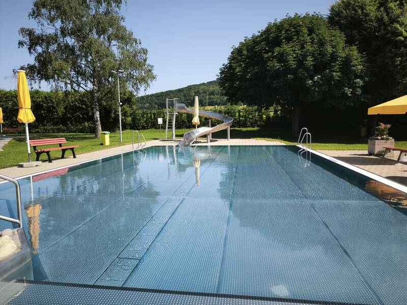 Ein Schwimmbecken im Freien mit einer kleinen Rutsche, umgeben von Bäumen und Sonnenschirmen.