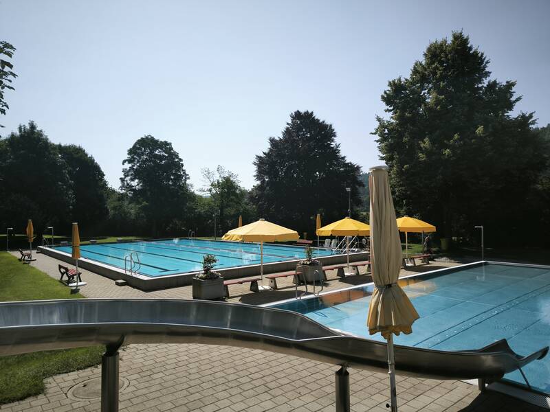 Ein Freibad mit einem großen rechteckigen Pool, gelben Sonnenschirmen, Rutsche und umgeben von Bäumen.