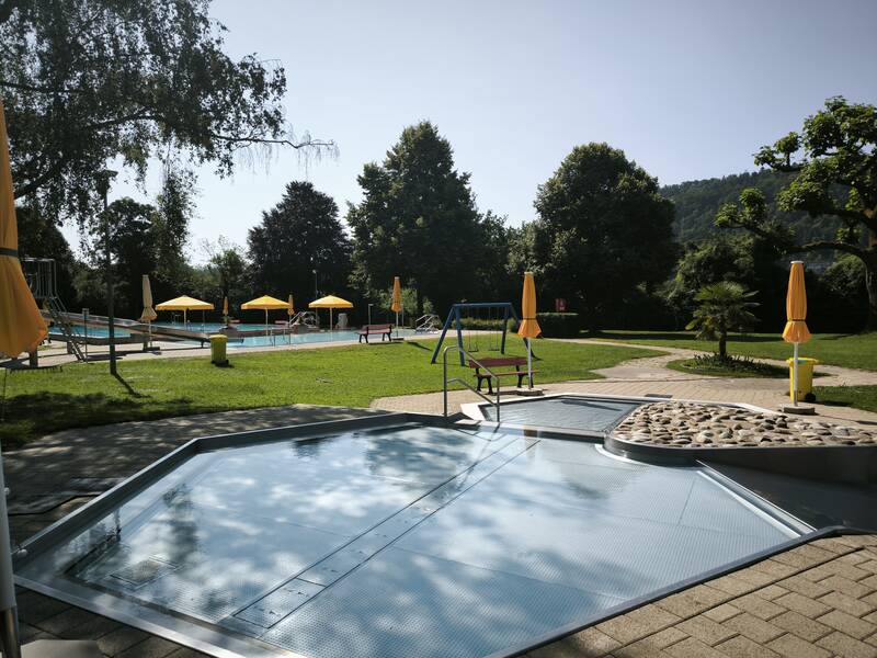 Ein Freibadbereich mit gelben Sonnenschirmen, Schwimmbecken und grünen Rasenflächen bei sonnigem Wetter.