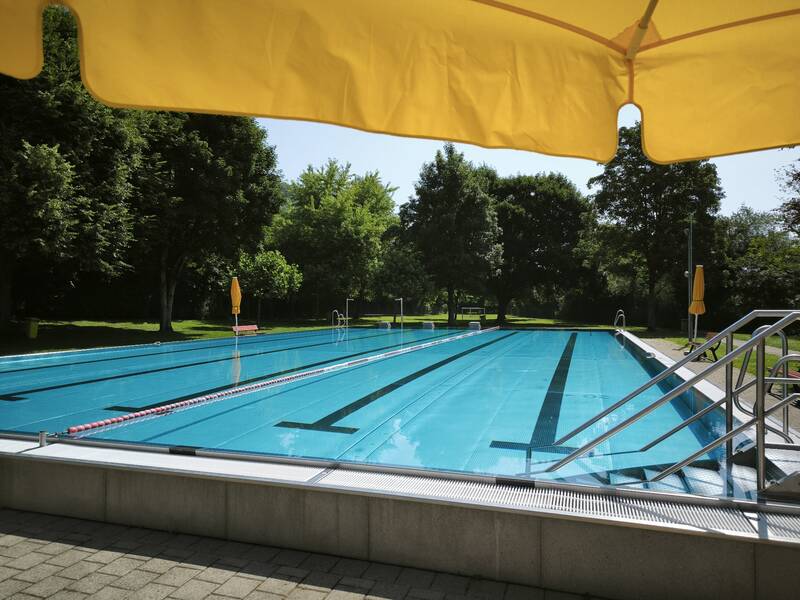 Ein Schwimmbad im Freien mit mehreren Bahnen, umgeben von Bäumen unter einem gelben Sonnenschirm.