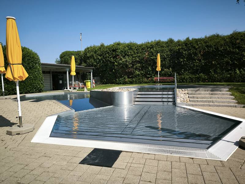 Eine Terrasse mit Treppen, die zu einem flachen Wasserbecken führen, ist von grüner Hecke umgeben, und mehrere gelbe Sonnenschirme sind aufgestellt.