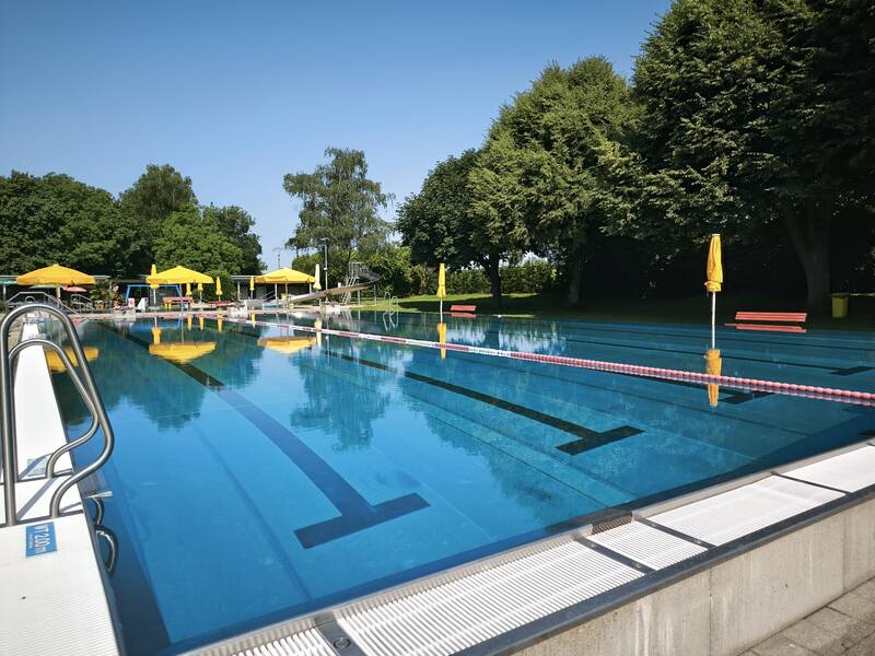 Ein großes Freibad mit mehreren gelben Sonnenschirmen und Bäumen im Hintergrund.