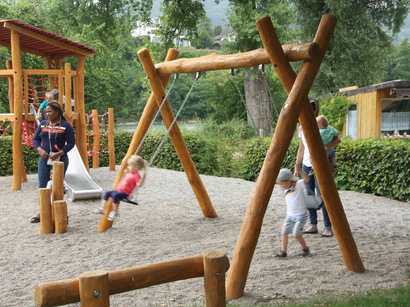 Auf einem Spielplatz mit Holzspielgeräten schaukeln Kinder, während ein Erwachsener in der Nähe steht und ein anderes Kind mit einem Erwachsenen im Hintergrund spielt.
