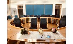 Ein runder Konferenzraum mit einem großen Holztisch, darauf Blumenarrangements und ein Hochzeitsbuch vor leeren Stühlen.