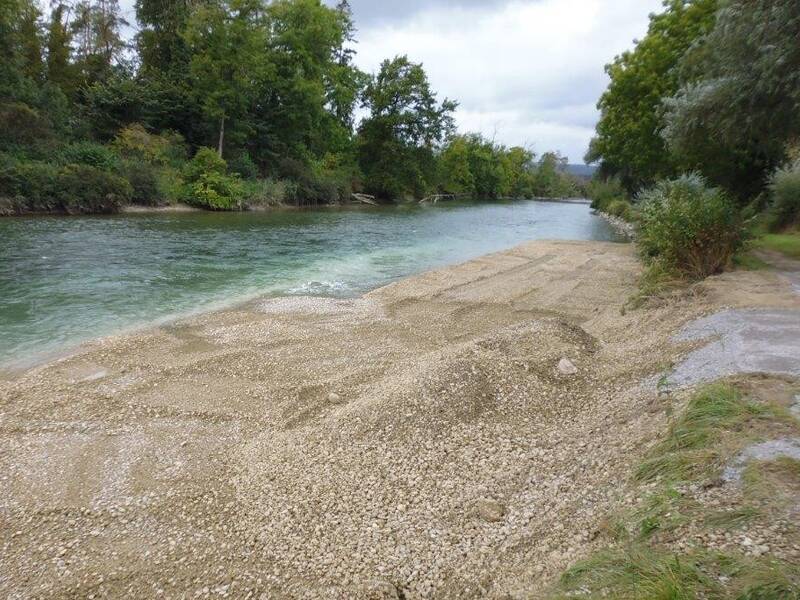 Ein Fluss fließt entlang einer kiesbedeckten Uferzone, umgeben von üppigem grünem Laub.