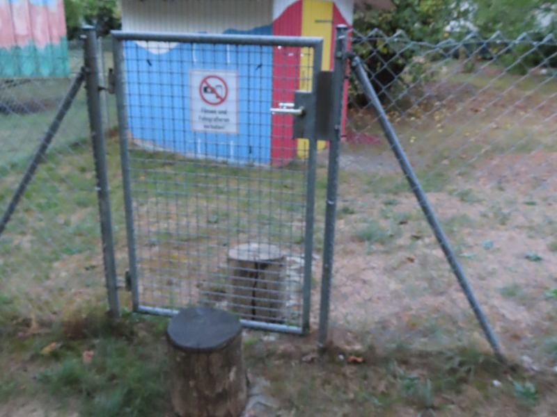 Ein kleiner bunt bemalter Schuppen steht hinter einem Drahtzaun mit einer Gartentür, die ein Verbotsschild trägt.