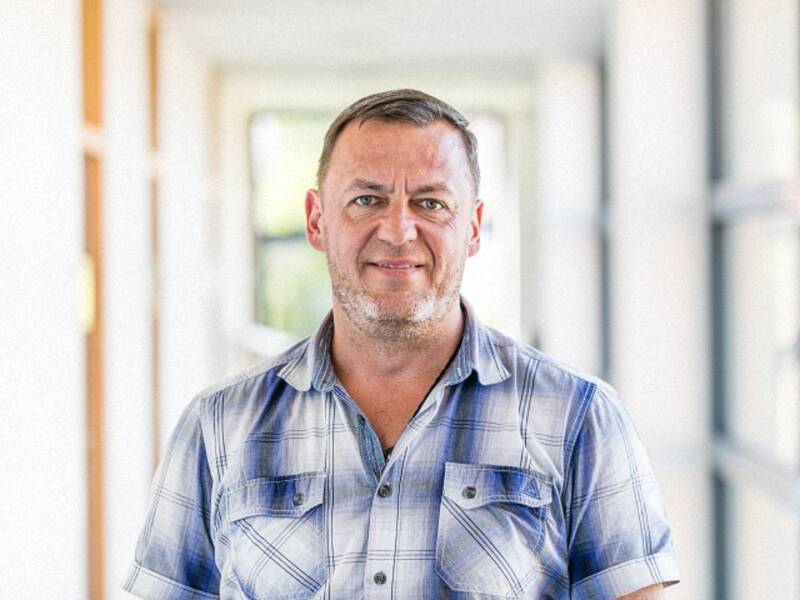 Mann mit kurzen Haaren und kariertem weiß-blauem Hemd steht in einem langen hellen Flur.