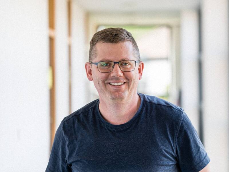 Mann mit kurzen Haaren, Brille und schwarzem T-Shirt steht in einem langen hellen Flur
