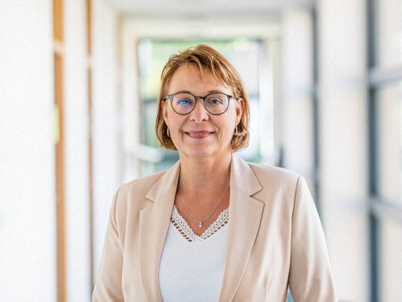 Frau mit kurzem braunem Haar, Brille und weißen Shirt steht in einem langen Hellen Flur