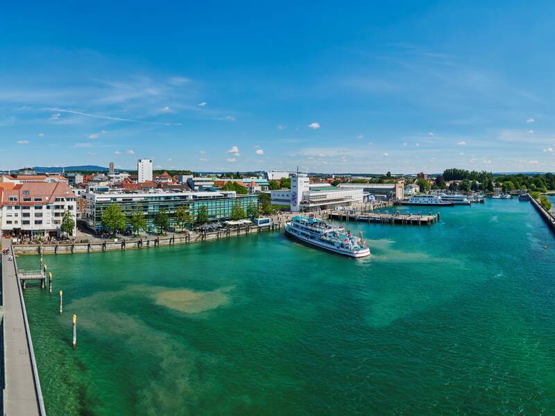Eine Panoramaaufnahme zeigt eine Küstenstadt an einem sonnigen Tag, umgeben von klarem, blauem Wasser und einem Hafen mit einem Schiff.